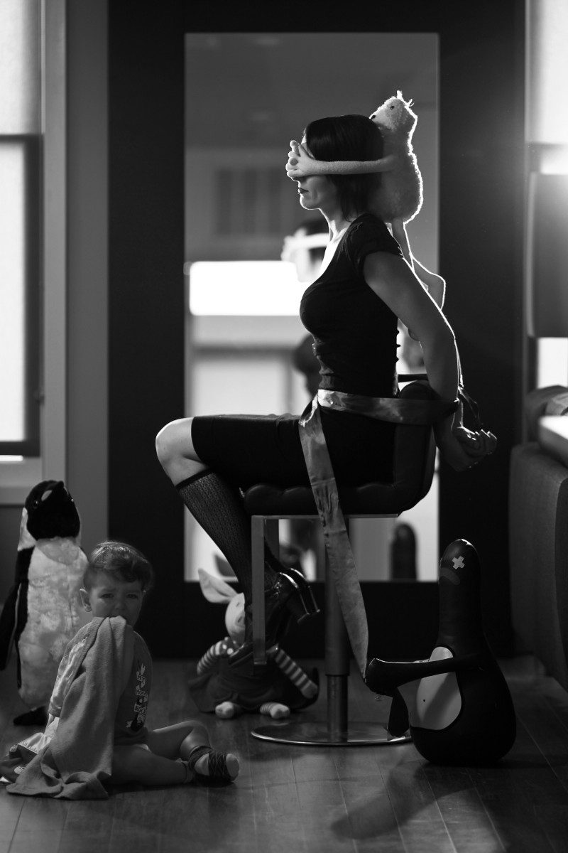 Woman in a business dress, tied to a chair with her eyes blindfolded with a stuffie. It appears as though the toys have captured her. On the floor to her left, a toddler is holding on to his blankie, upset.